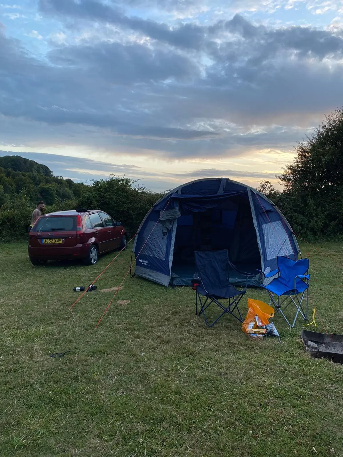 Photo of A blue camping tent set up next to a parked red car in a grassy area during sunset. | Wander Sober
