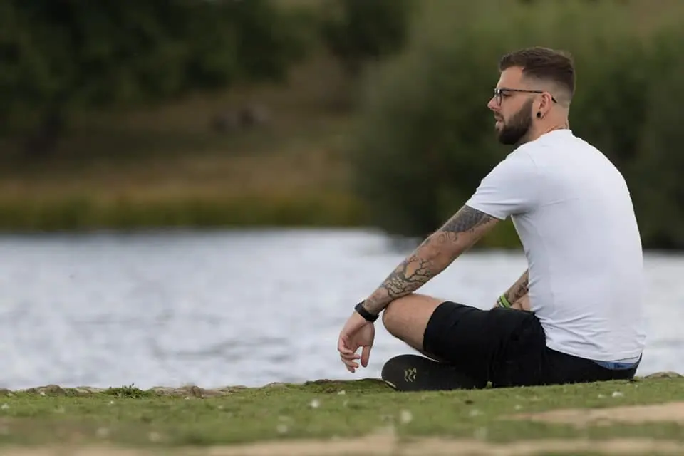 Photo of Man with tattoos sitting on the grass by a lake, wearing a white t-shirt and shorts. | Wander Sober