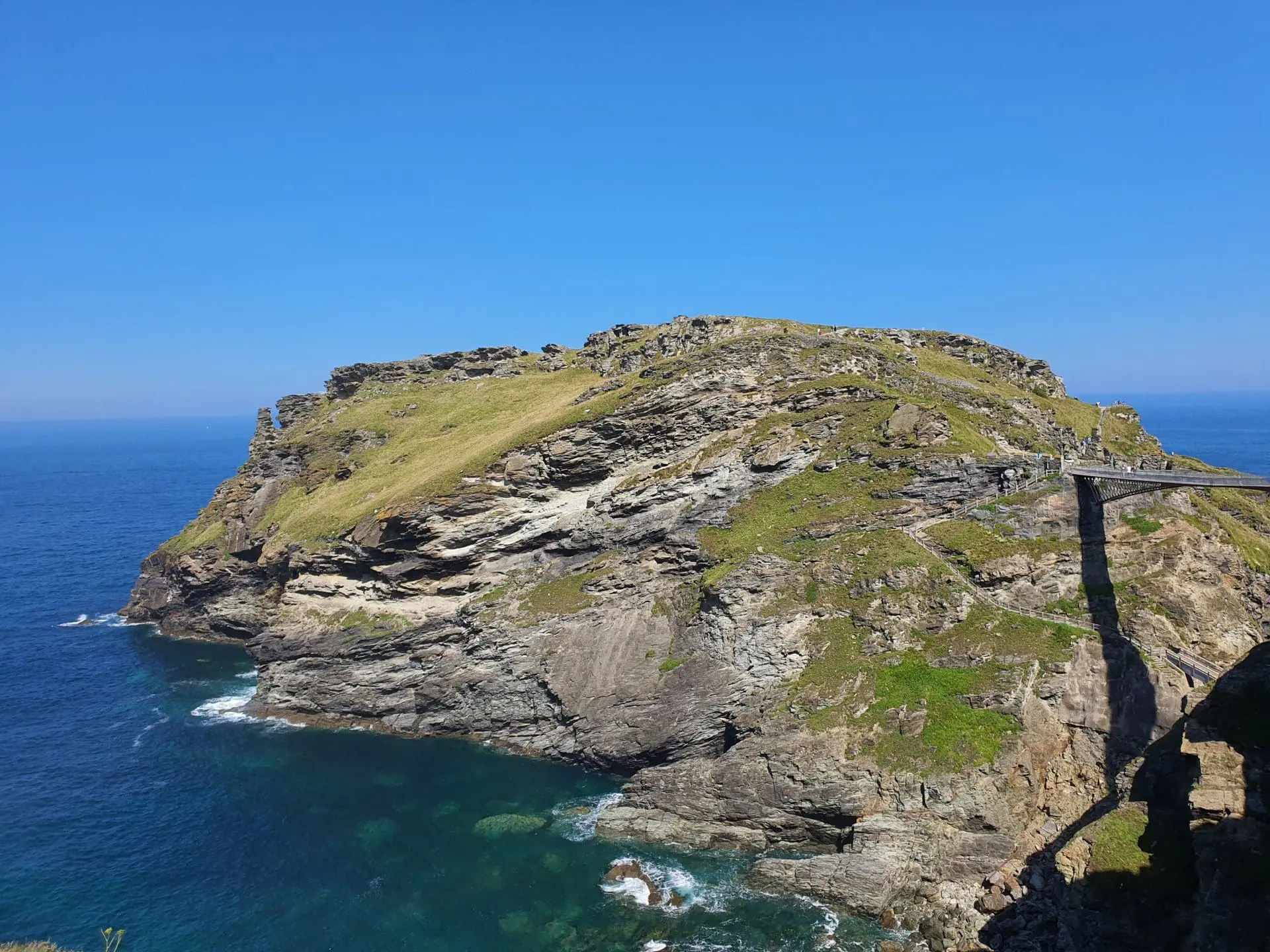 Photo of Rocky coastal formation with greenery on top and clear blue water surrounding it. | Wander Sober