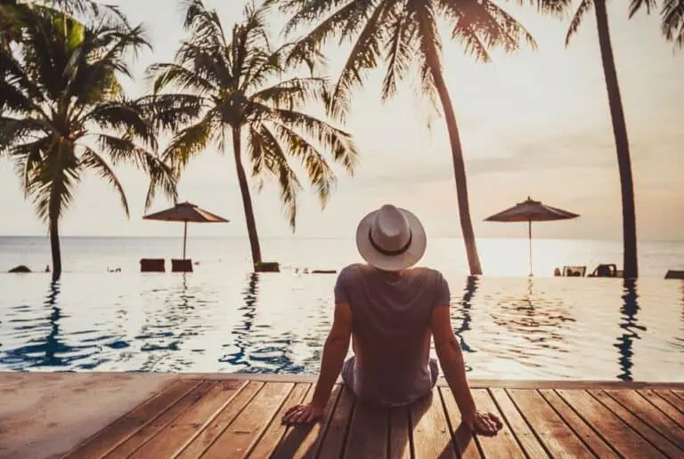 Photo of Individual wearing a hat sitting by an infinity pool with palm trees at sunset. | Wander Sober