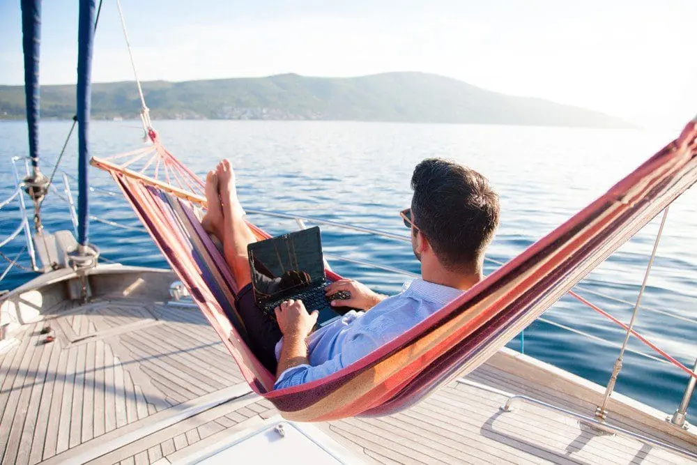Photo of Man using a laptop while lounging in a hammock on a boat with water in the background. | Wander Sober