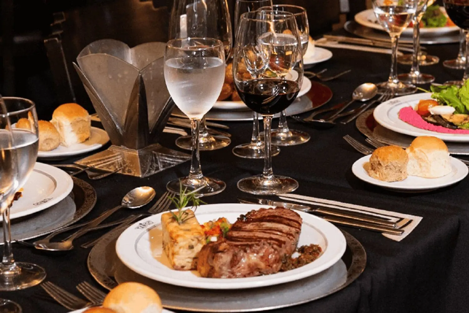 Photo of A dining table set with steak, wine glasses, and rolls on black tablecloth. | Wander Sober