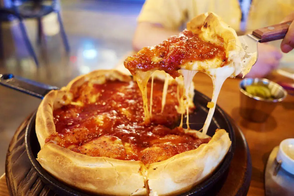 A slice of deep dish pizza being lifted from a pan, showing melted cheese and tomato sauce.