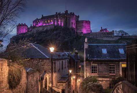 Photo of Edinburgh Castle illuminated in purple light at dusk, situated on a rocky hill. | Wander Sober