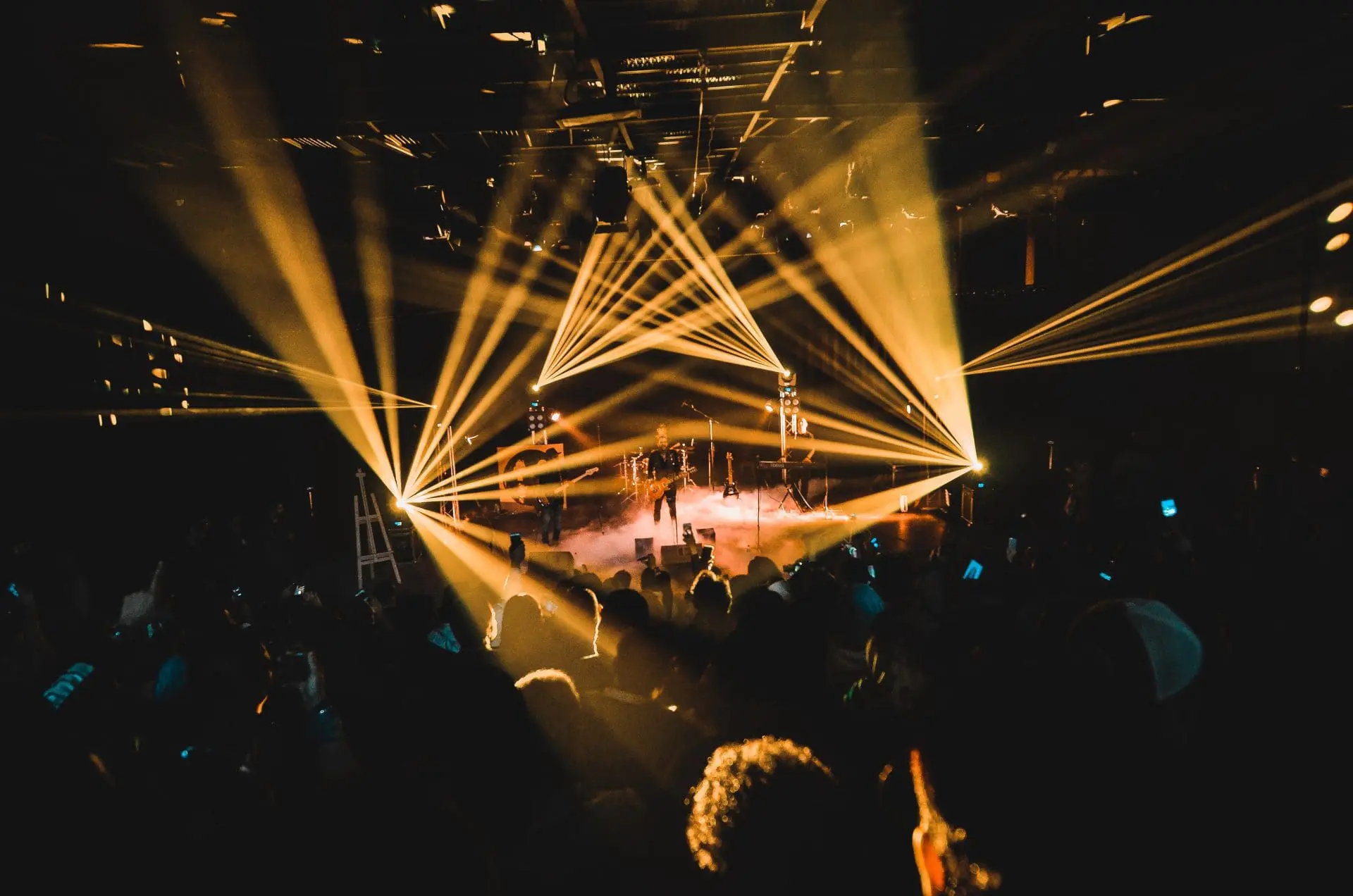 Concert stage illuminated by beams of light with an audience in the foreground.