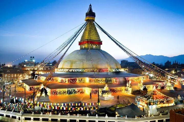 Photo of Boudhanath Stupa illuminated at dusk with prayer flags and lights in Kathmandu, Nepal. | Wander Sober