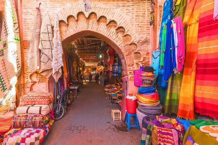 Photo of Narrow market street in Marrakech lined with vibrant textiles and goods for sale. | Wander Sober