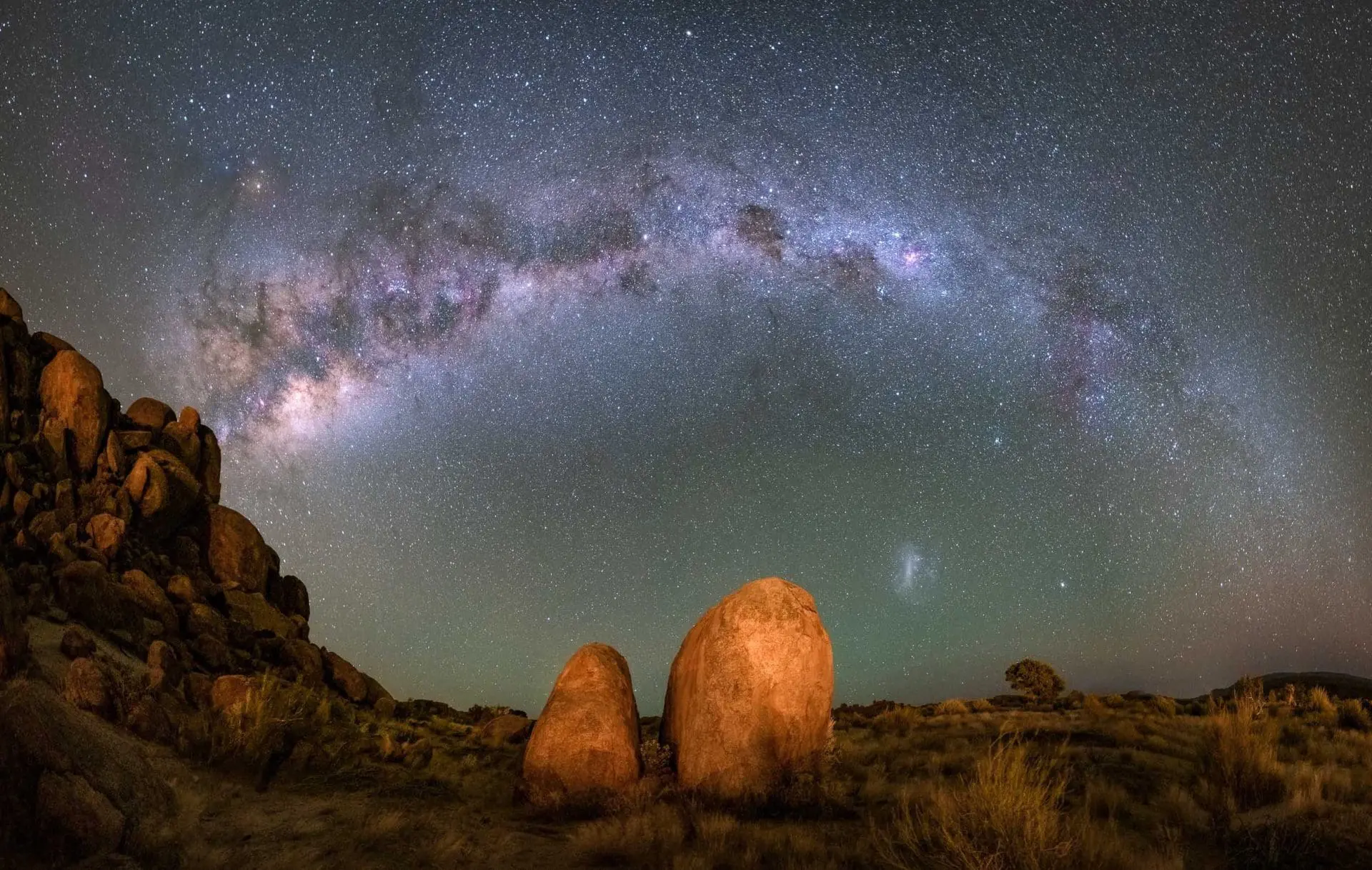 Panoramic view of the Milky Way galaxy arching over two large rocks in a natural setting.