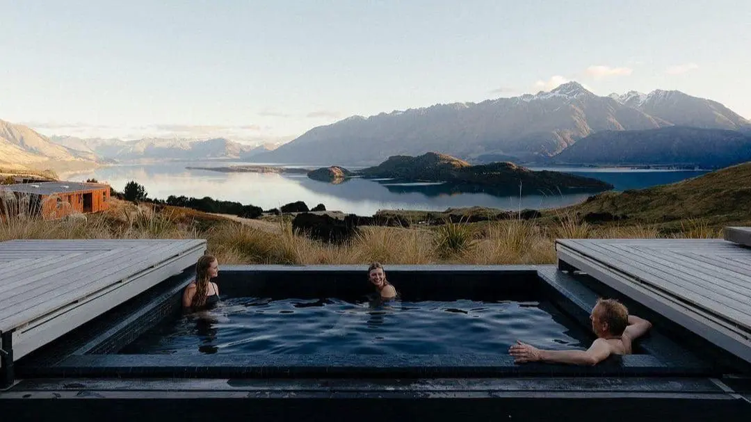 Photo of Three people relaxing in an outdoor hot tub with mountains and a lake in the background. | Wander Sober