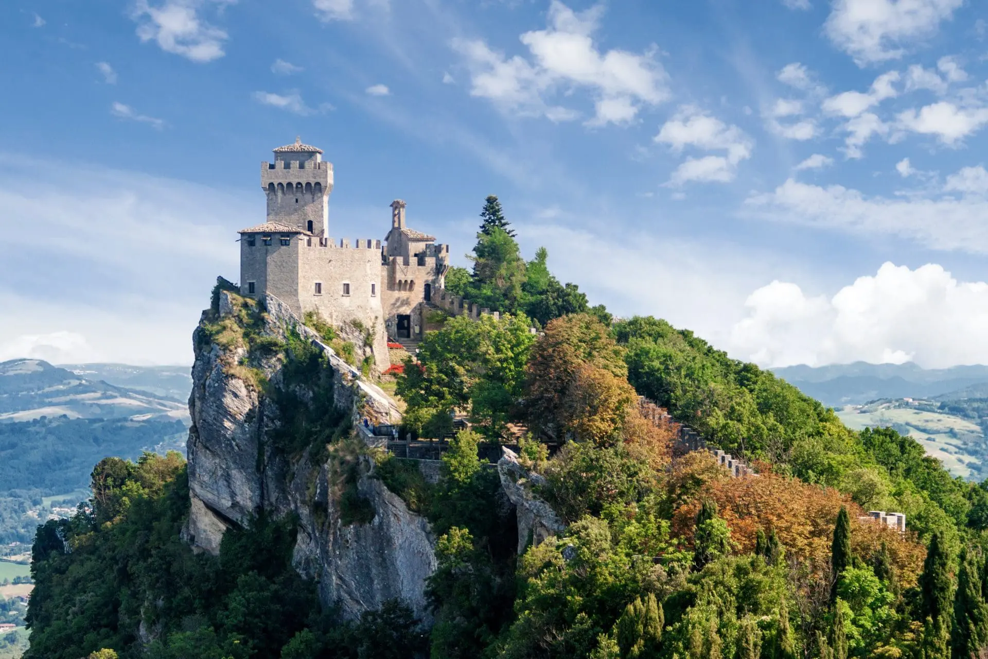 Photo of A stone castle situated on a rocky hill surrounded by greenery and trees under a blue sky. | Wander Sober