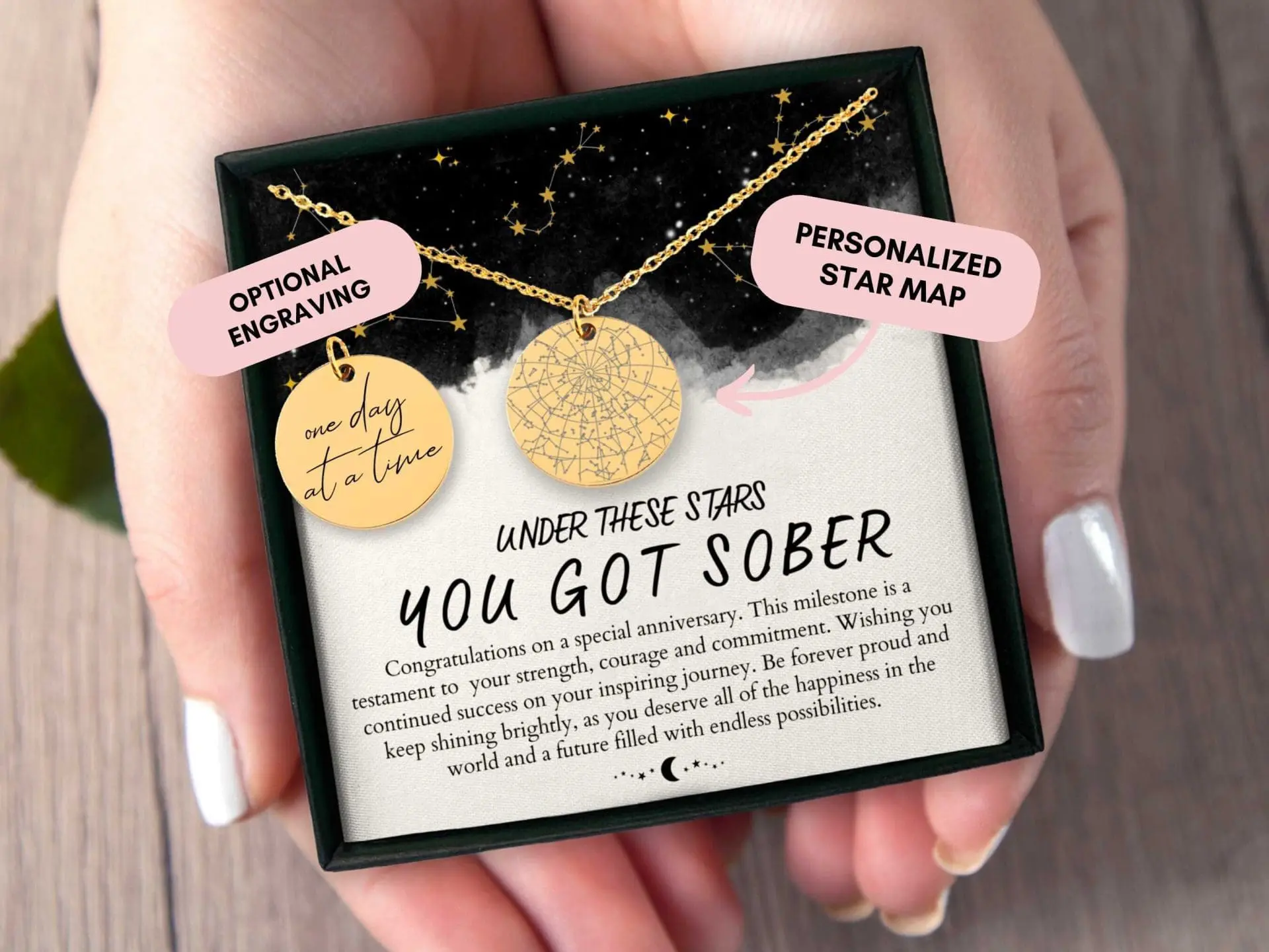 Photo of Gold necklace featuring a star map pendant and an engraved disc pendant in a gift box held by a hand. | Wander Sober