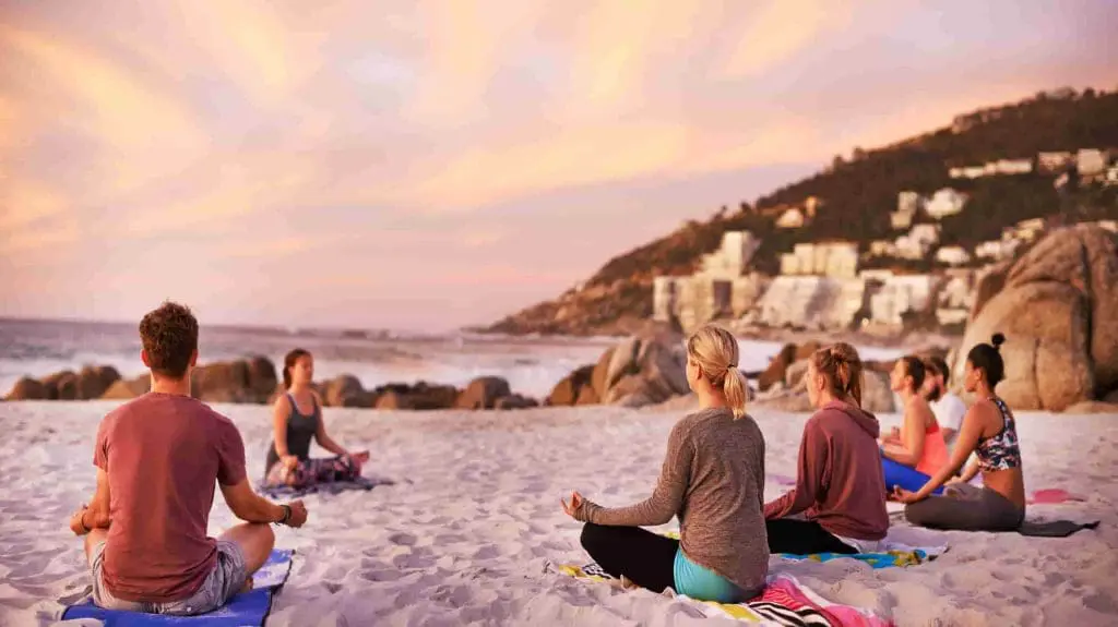 Photo of A group of six individuals practicing meditation on a sandy beach during sunset. | Wander Sober