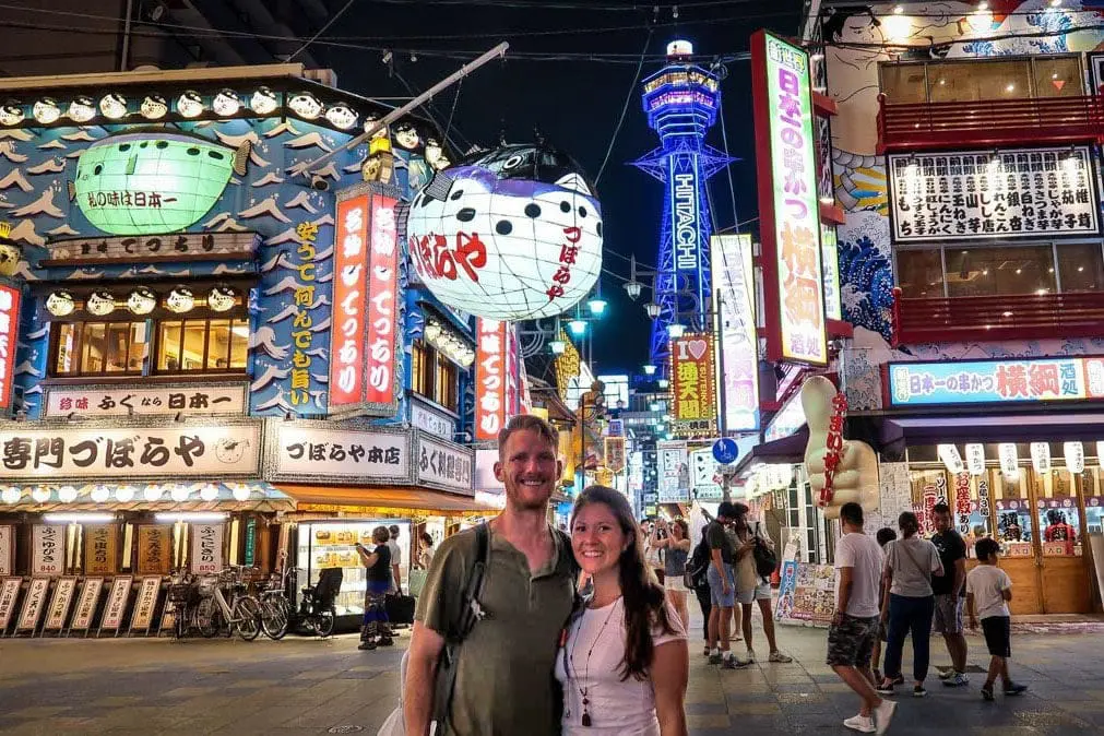 Photo of A couple standing together in a brightly lit street filled with neon signs and colorful storefronts at night. | Wander Sober