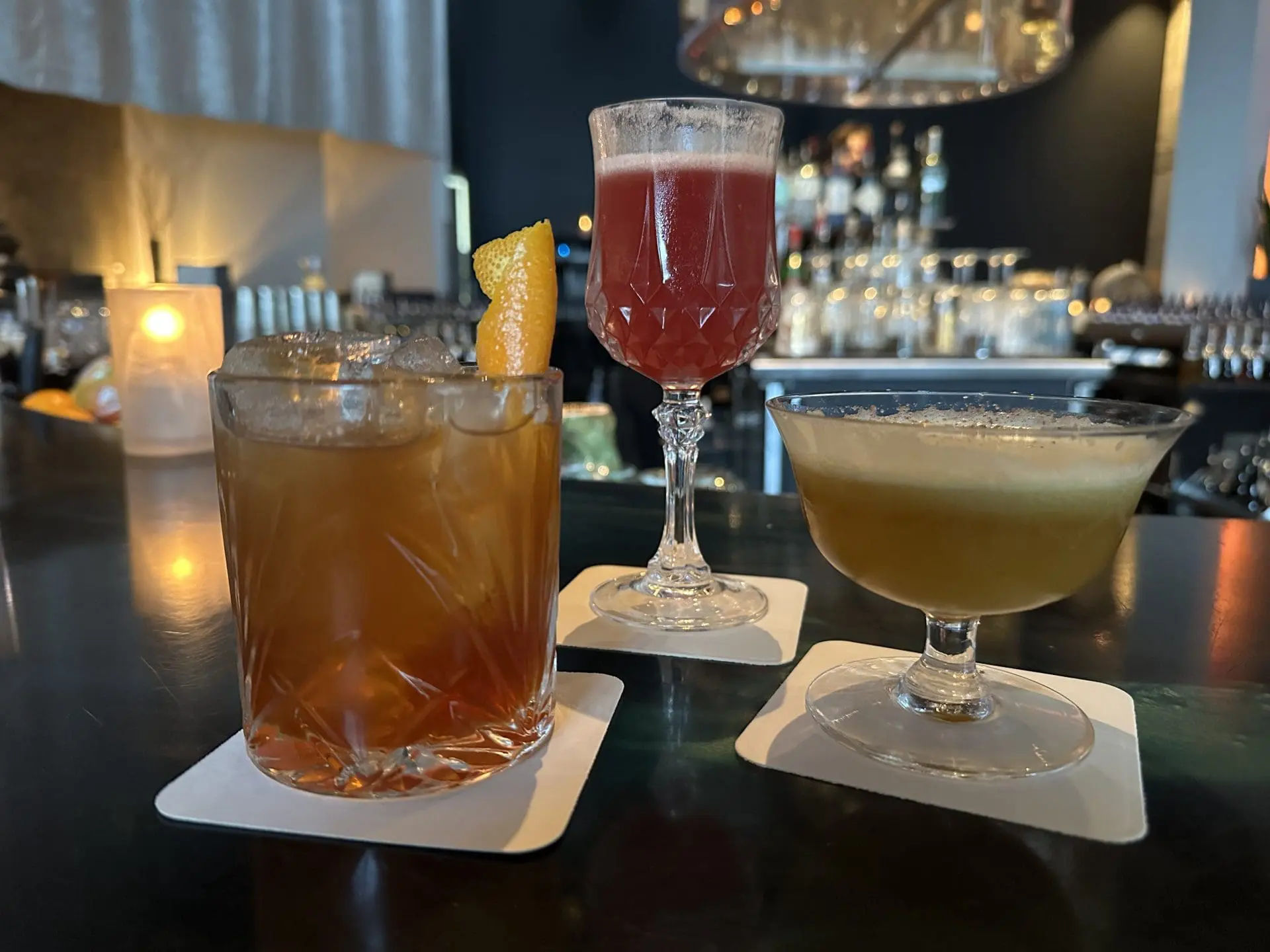 Photo of Three cocktails displayed on a bar counter, including a brown drink with an orange garnish and two other colorful drinks. | Wander Sober