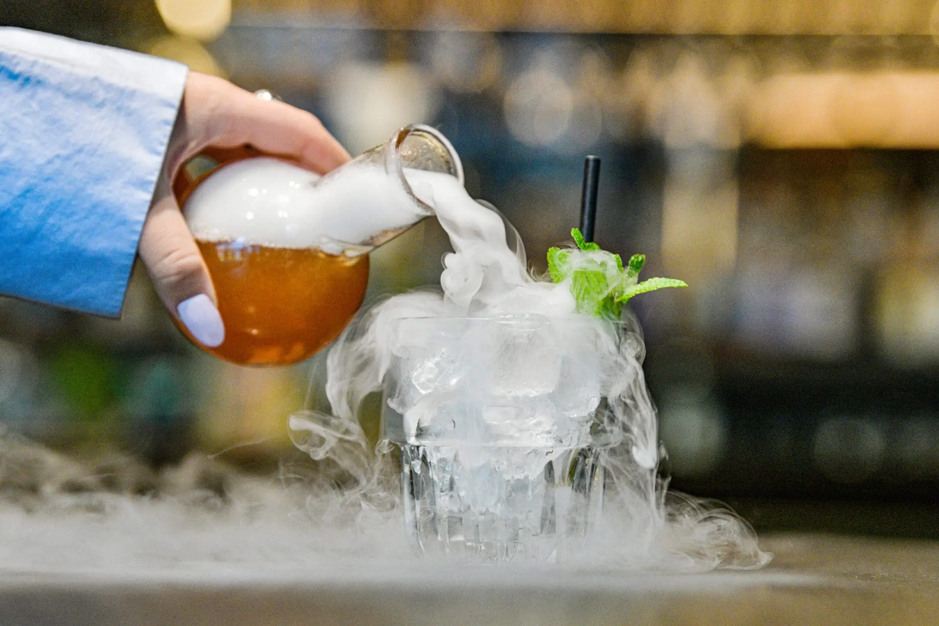 Photo of A hand pouring a drink from a flask into a glass filled with ice and smoke, garnished with mint. | Wander Sober
