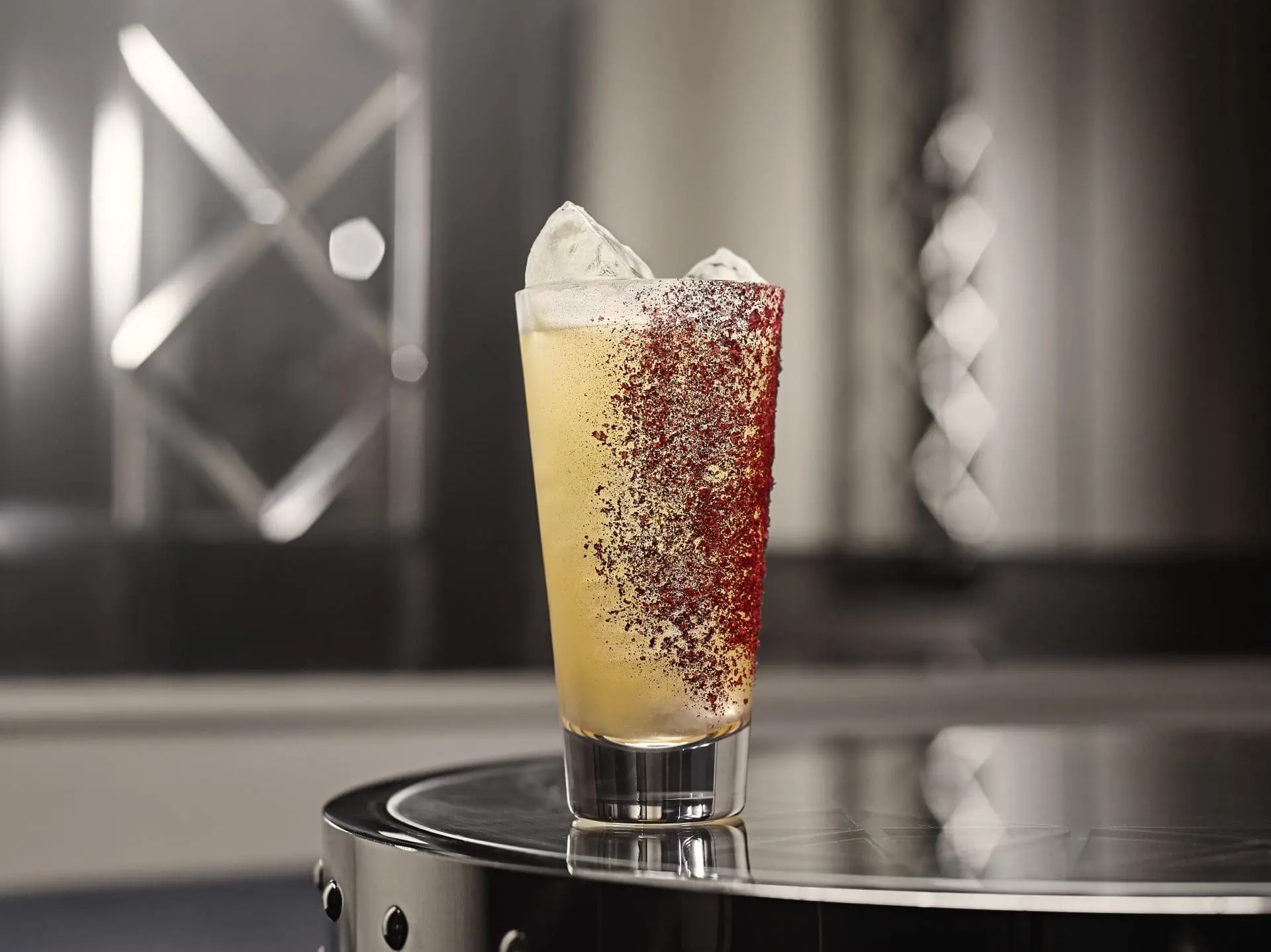 Photo of A tall glass of cocktail with ice and a red rim, placed on a reflective surface. | Wander Sober