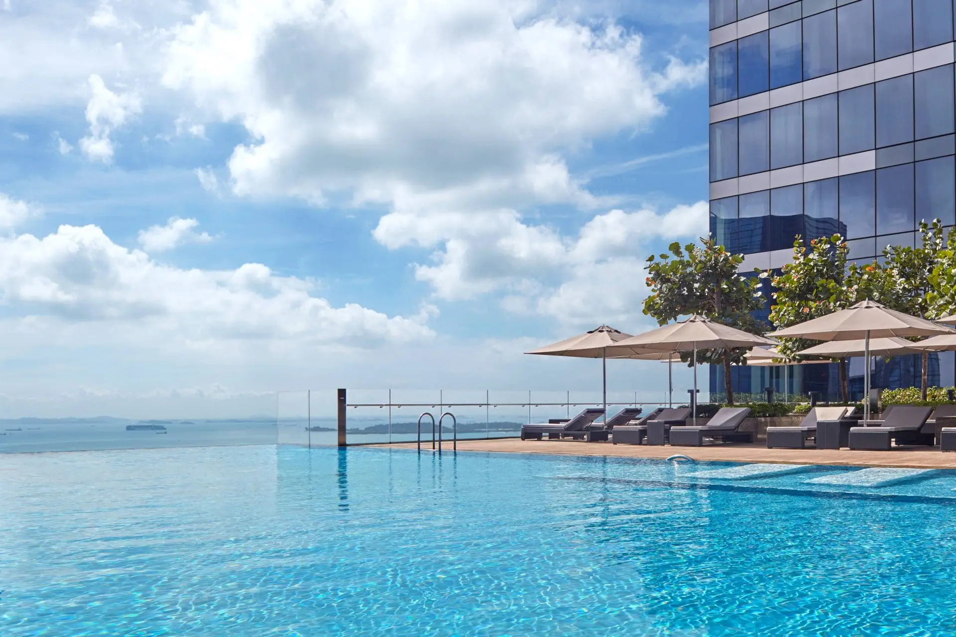 Photo of Infinity pool with lounge chairs and umbrellas beside a modern building under a cloudy sky. | Wander Sober