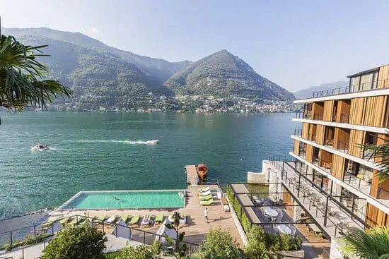 Photo of View of a modern hotel with a swimming pool beside a lake and mountains in the background. | Wander Sober