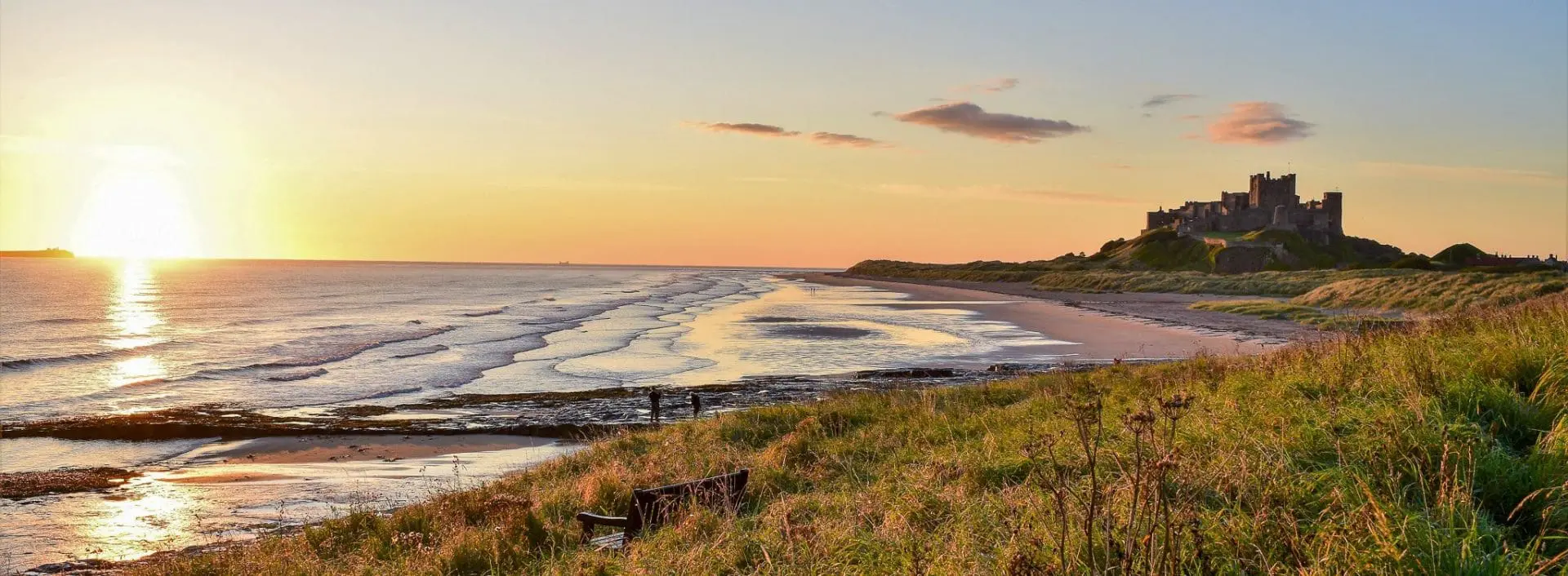 Photo of Bamburgh Castle situated on a hill overlooking a beach at sunset with gentle waves in the foreground. | Wander Sober