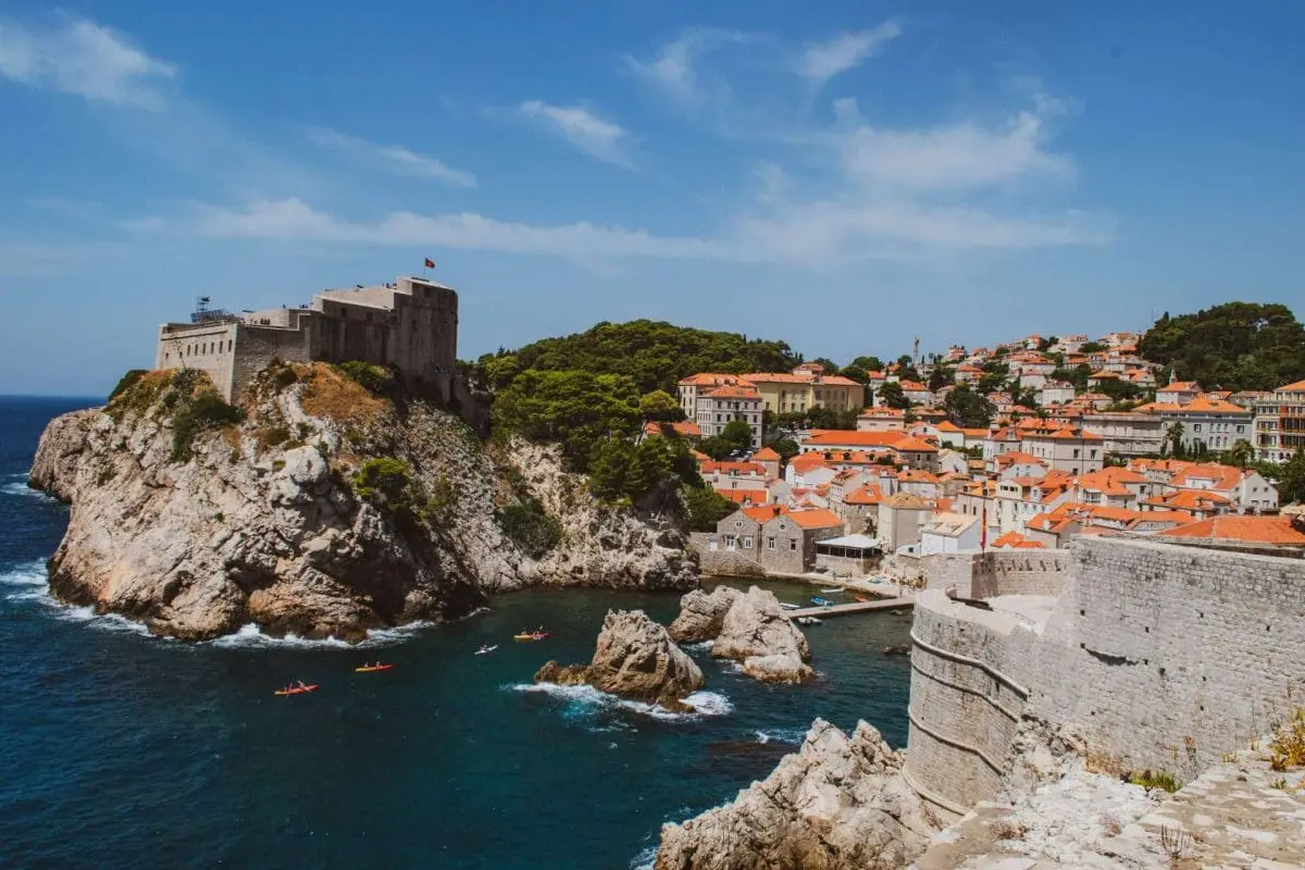 Photo of A coastal view of Dubrovnik featuring a fort on a rocky outcrop and a town with red-roofed buildings. | Wander Sober