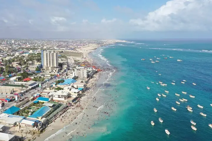 Photo of Aerial view of a coastal city featuring a beach, buildings, and several boats in the water. | Wander Sober