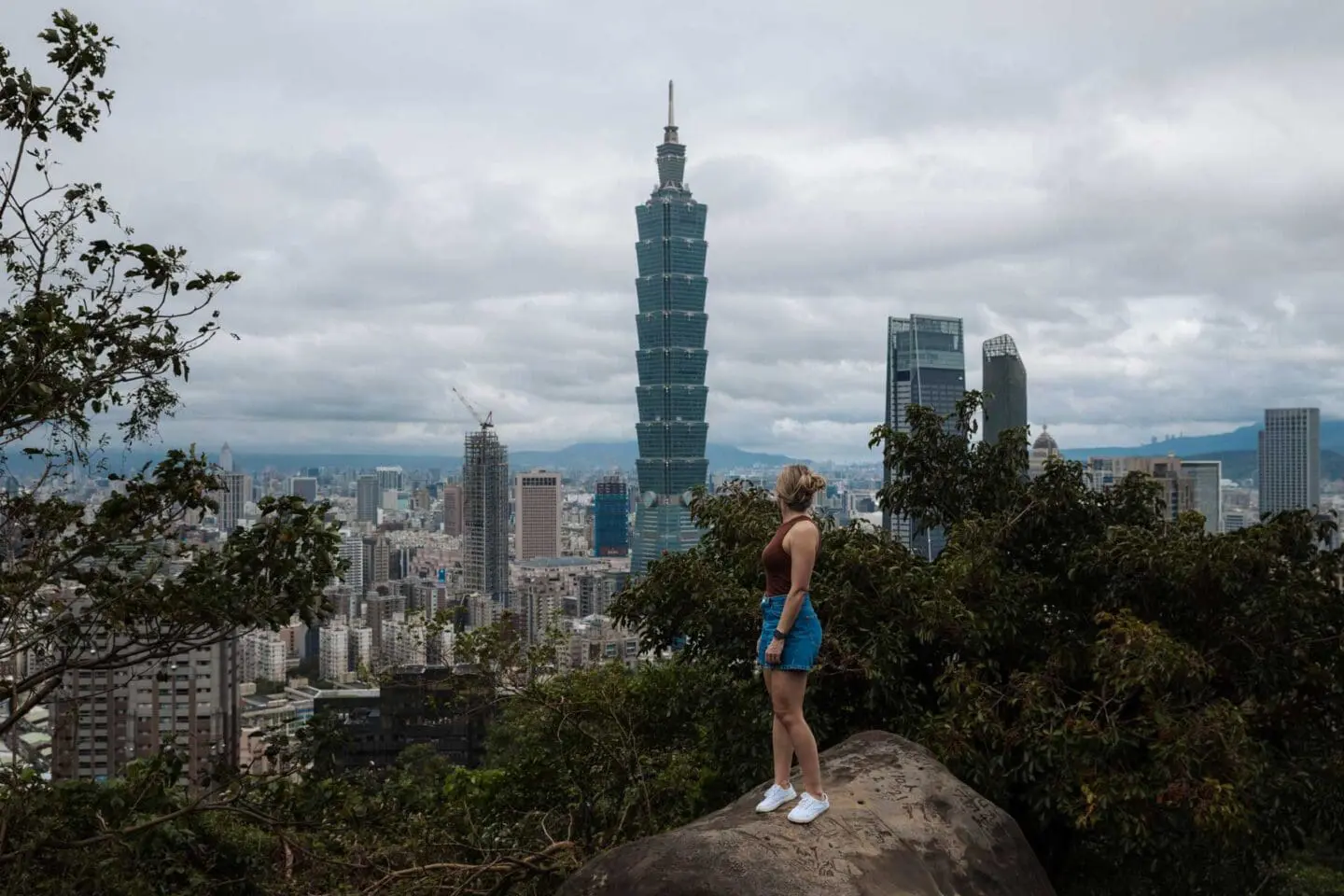 Photo of Individual standing on a rock with Taipei 101 skyscraper visible in the background amidst a cityscape. | Wander Sober