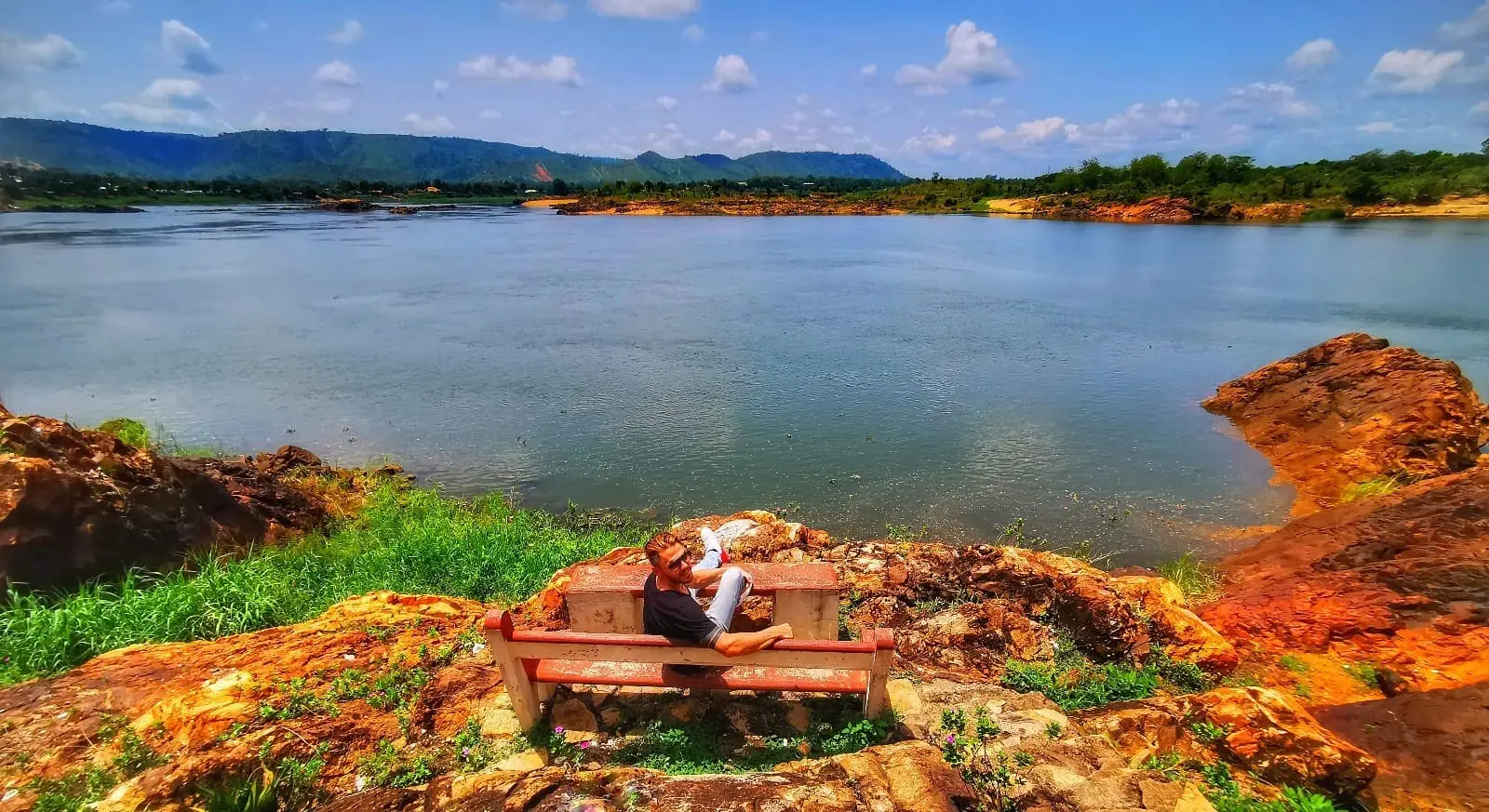 Photo of Individual sitting on a wooden bench by a calm lake surrounded by greenery and rocky terrain. | Wander Sober