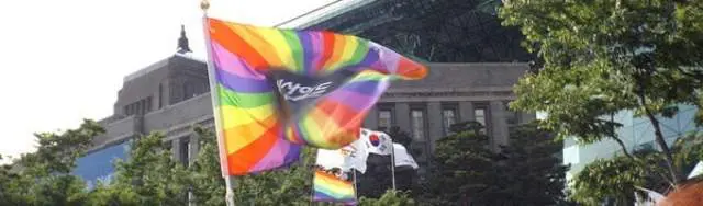 Photo of Rainbow pride flag waving in the air during a public event with buildings in the background. | Wander Sober
