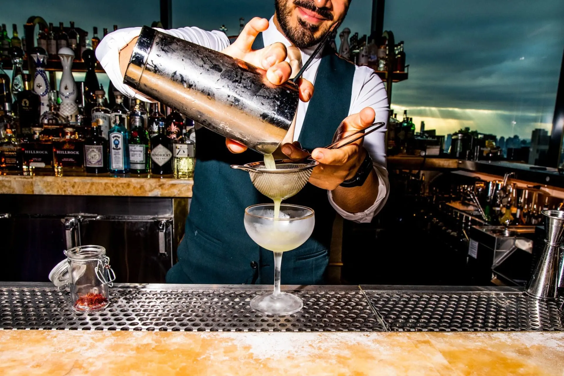 Photo of Bartender using a shaker to pour a cocktail into a glass with a strainer at a bar. | Wander Sober