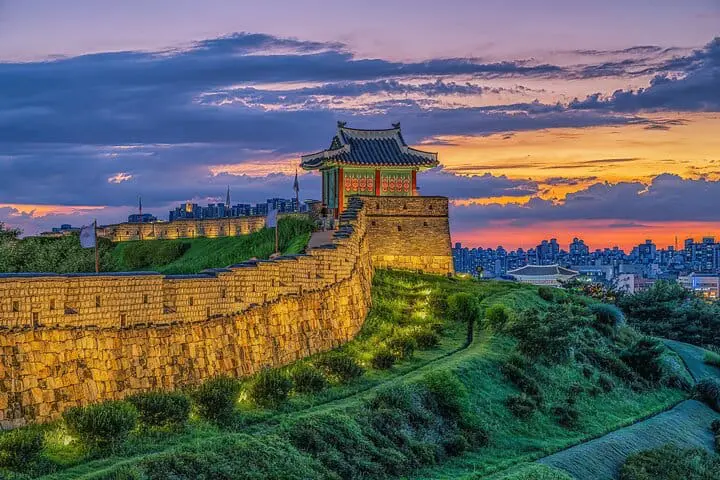 Photo of A historic fortress wall with a traditional pavilion at sunset, surrounded by greenery and city skyline. | Wander Sober
