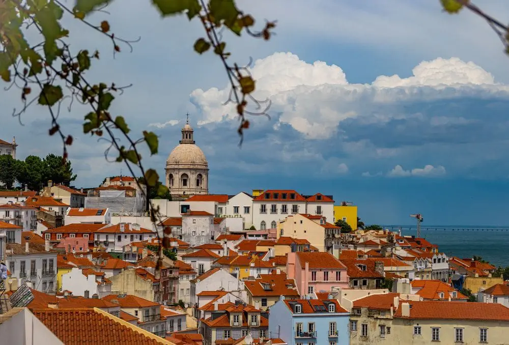 Photo of View of Lisbon cityscape featuring buildings with red roofs and a prominent dome structure under a cloudy sky. | Wander Sober