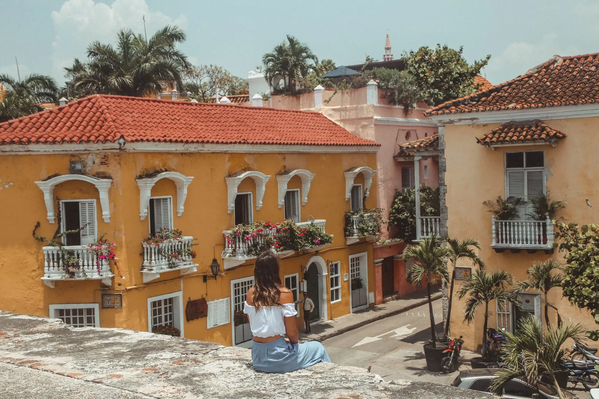 Photo of A woman in a white top and blue skirt sitting on a wall overlooking colorful buildings in Cartagena. | Wander Sober