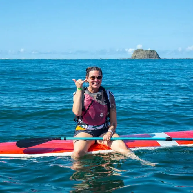 Photo of Individual sitting on a colorful paddleboard in the ocean, making a hand gesture. | Wander Sober