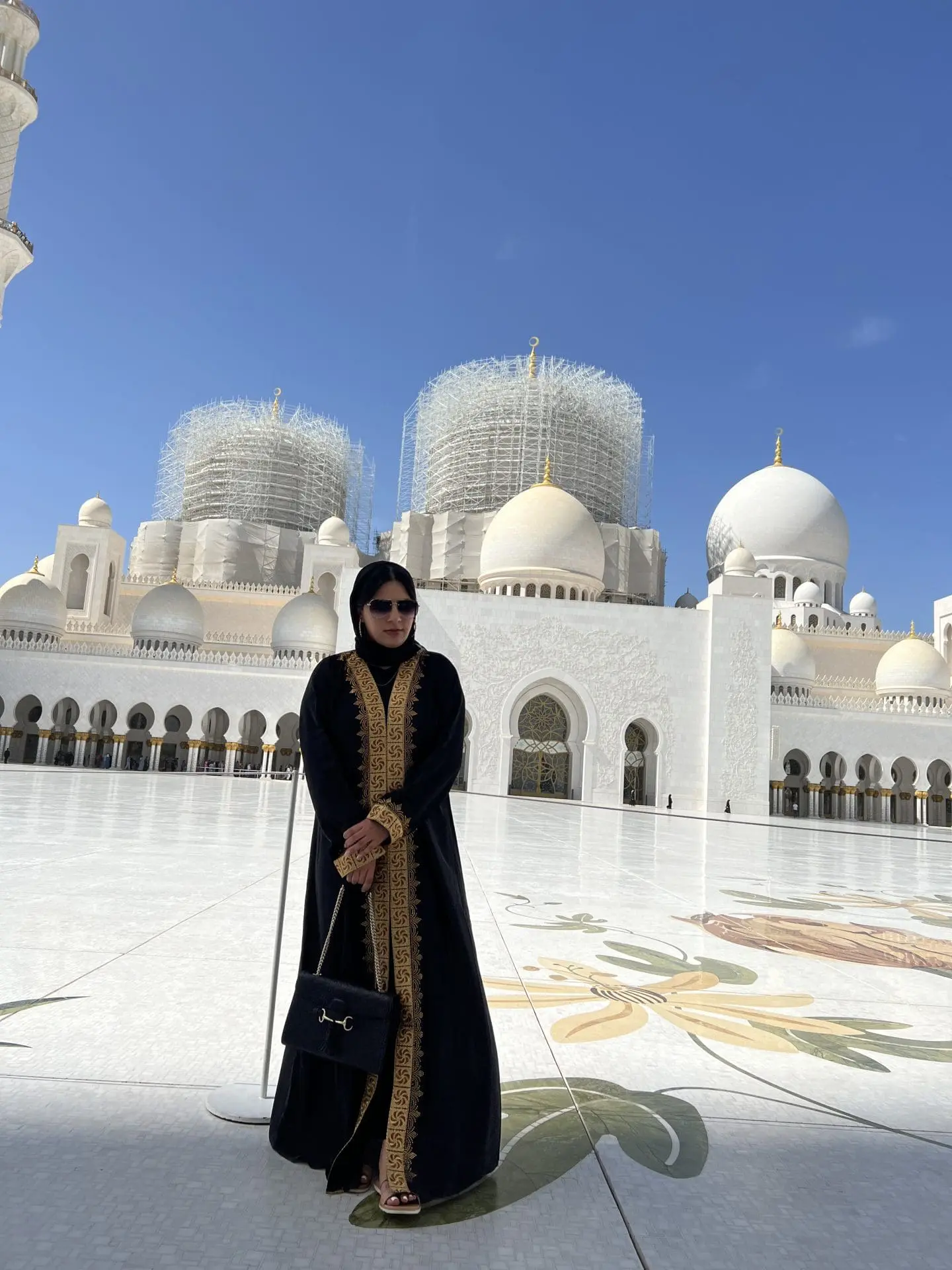 Photo of Woman wearing a black abaya with gold embroidery standing in front of a mosque under construction. | Wander Sober