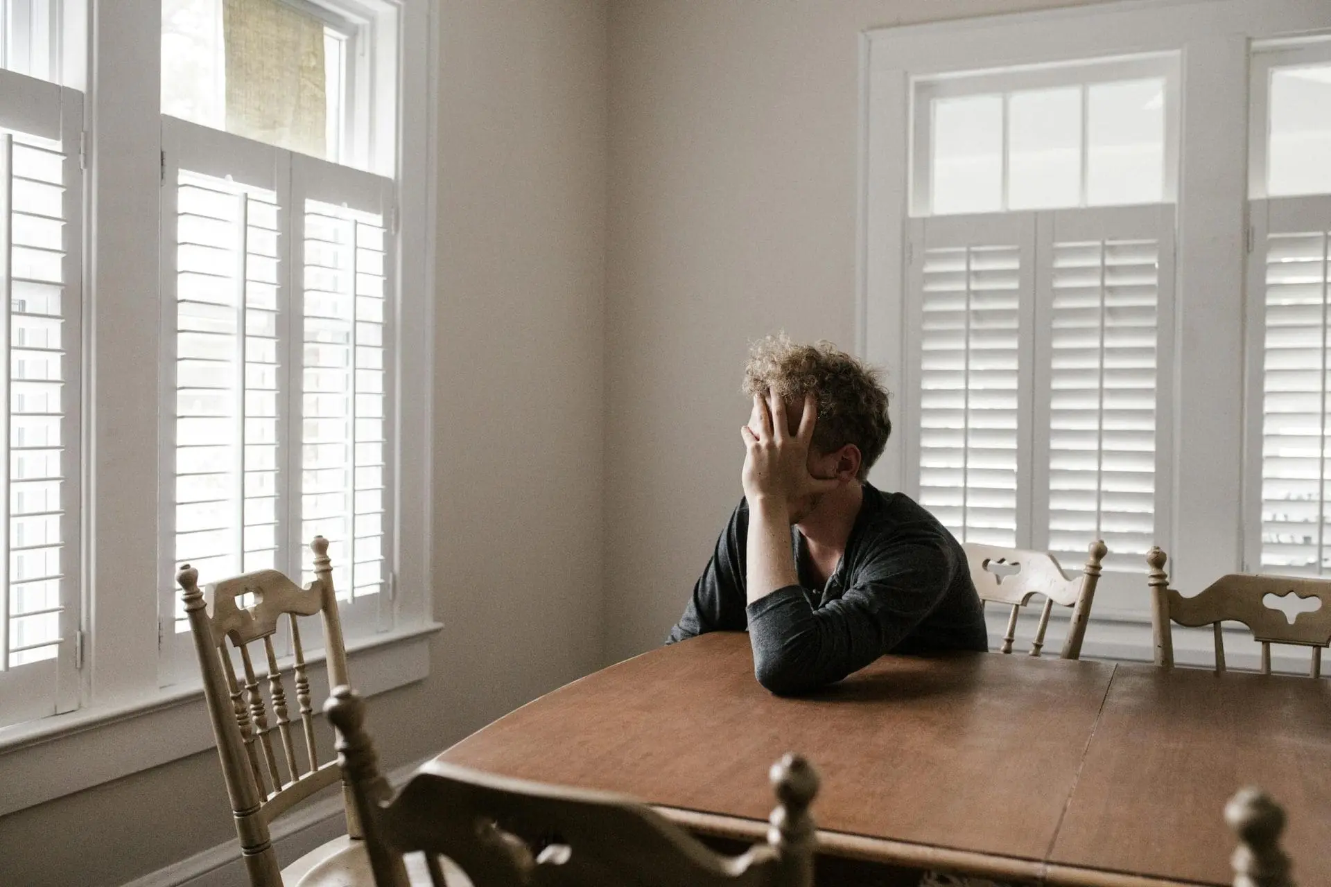Individual sitting at a wooden table with one hand covering their face, looking contemplative.