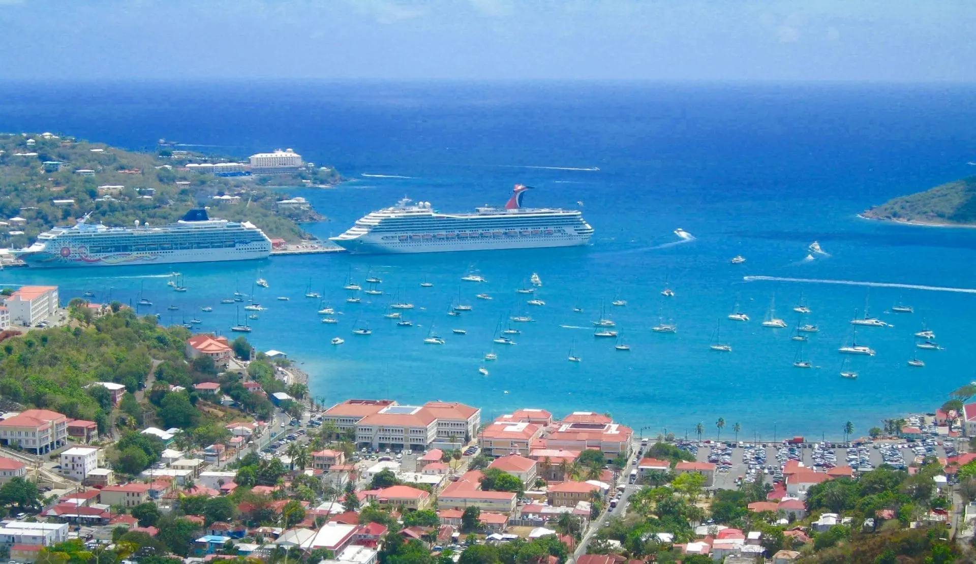 Aerial view of a tropical harbor featuring two cruise ships and several sailboats in clear blue waters.