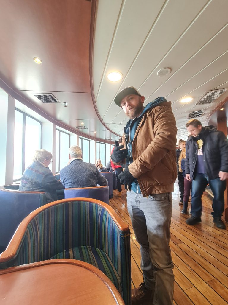 That Time I Thought I Saw French John McClane on a Ferry to France 3 Man posing with a peace sign in a cozy ferry lounge, surrounded by passengers enjoying the view.