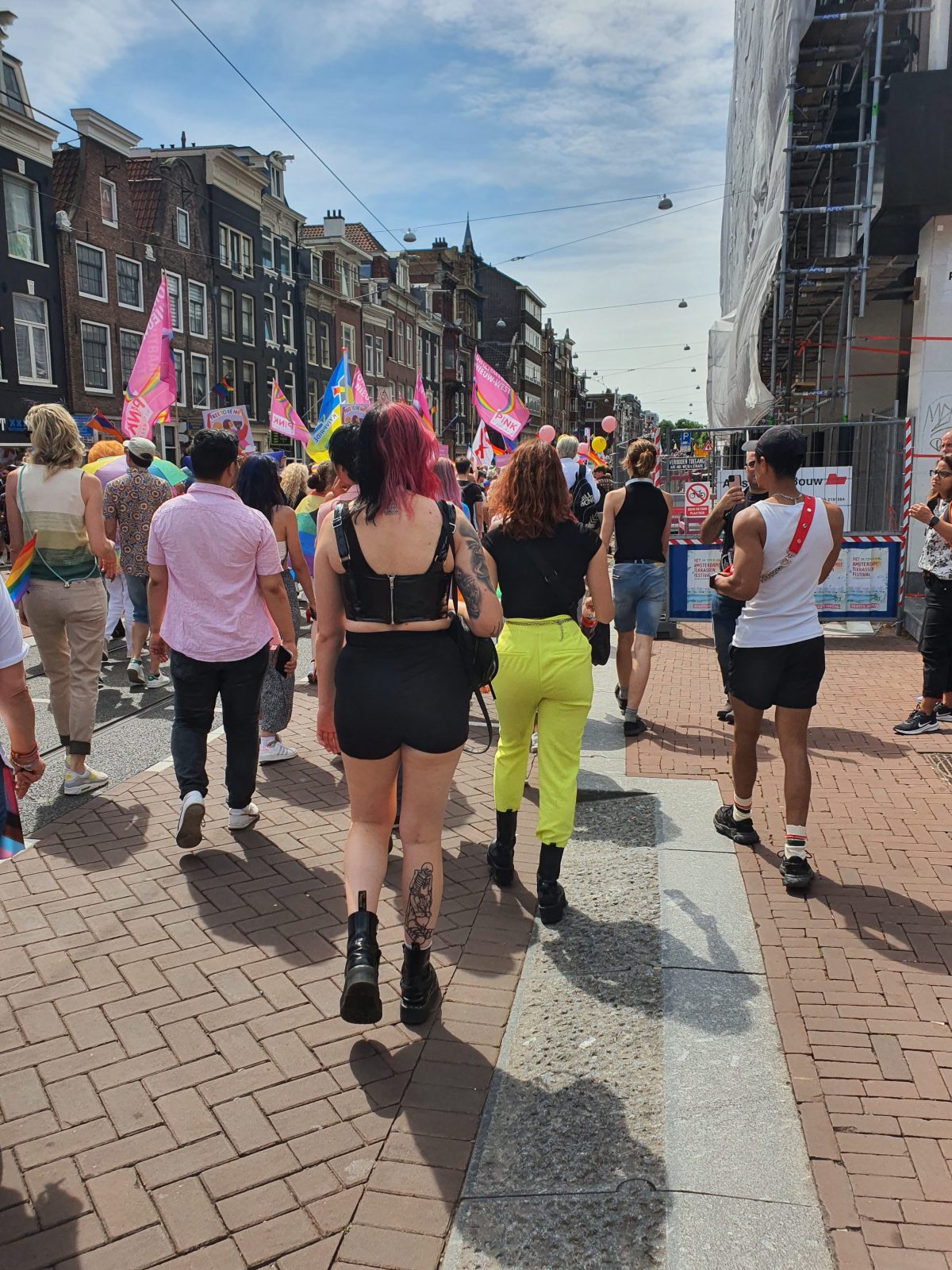 The Unforgettable Joy of Amsterdam Pride: Love, Freedom, and a Whole Lot of Glitter 6 Sover Travel - A lively street scene unfolds on the sunlit streets of Malta, with people walking and some waving colorful flags, likely portraying a parade or festival. Participants wear vibrant attire. Historic buildings with gabled roofs and scaffolding are visible against a partly cloudy sky.