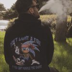 Sover Travel - A person sitting outdoors, exhaling vapor into the air. They wear a black hoodie featuring an image of an animated character with the text, DONT LIGHT A FIRE IF YOU CANT HANDLE THE SMOKE. Surrounding them are trees and grass.