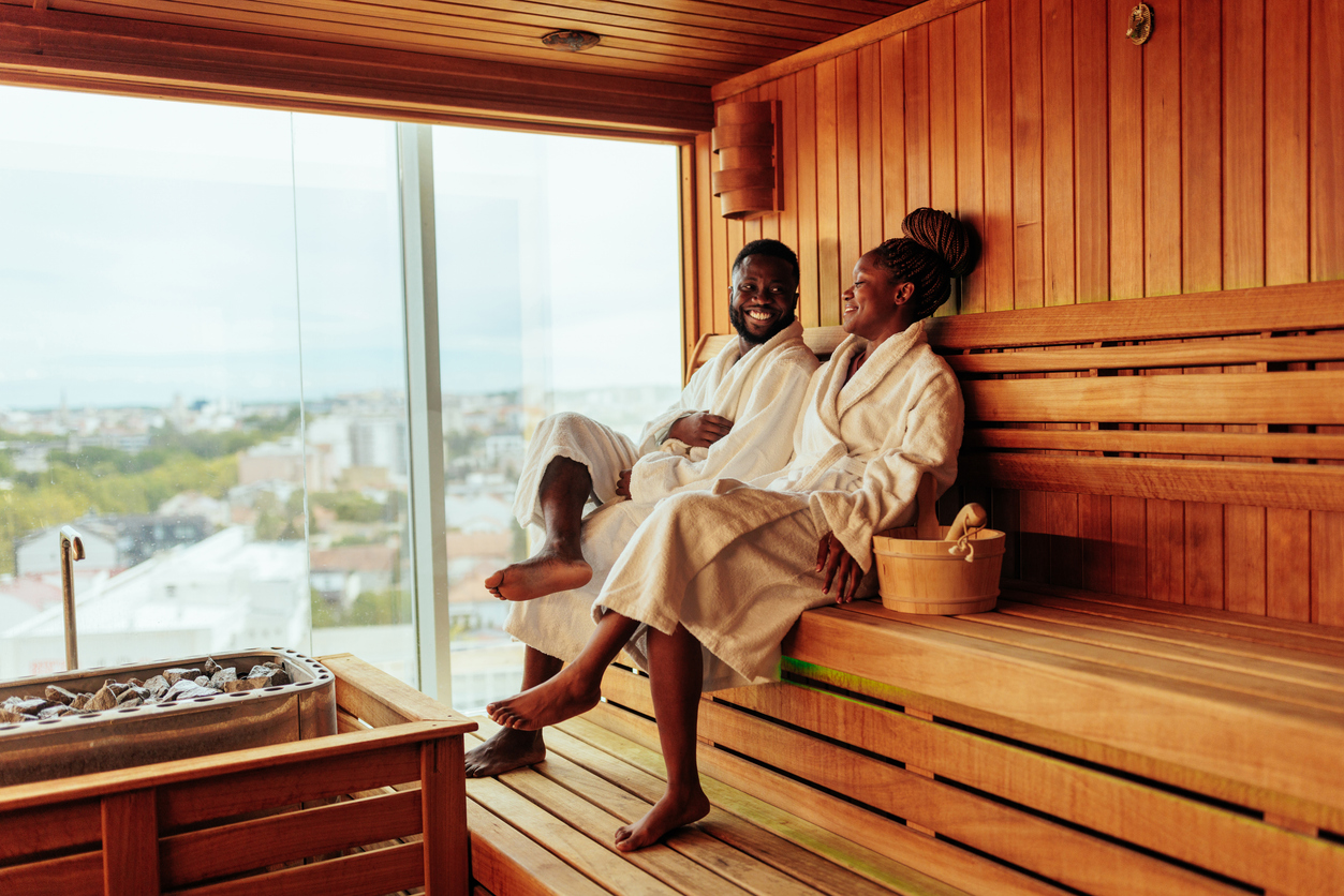 Alcohol-Free Travel Best Places: Discover Inspiring Destinations for Sober Explorers 4 Sover Travel - Two people in white robes sit smiling in a wooden sauna with a city view through a large glass window, embodying the spirit of sober explorers. A bucket with a ladle is placed beside them on the bench, adding to their alcohol-free travel adventure.