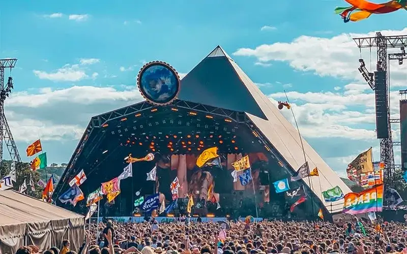 Photo of Pyramid stage at a music festival with a large crowd and various flags in the foreground. | Wander Sober