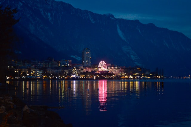 Montreux Jazz Festival: Lakeside Tunes, Swiss Culture, and NA Experiences - Finding Sober Joy Along Lake Geneva's Musical Shoreline 6 Sover Travel - A nighttime view of a lakeside town with a lit Ferris wheel reflecting in the water. Snow-capped mountains form a backdrop. The towns lights create a warm glow against the deep blue sky and lake.
