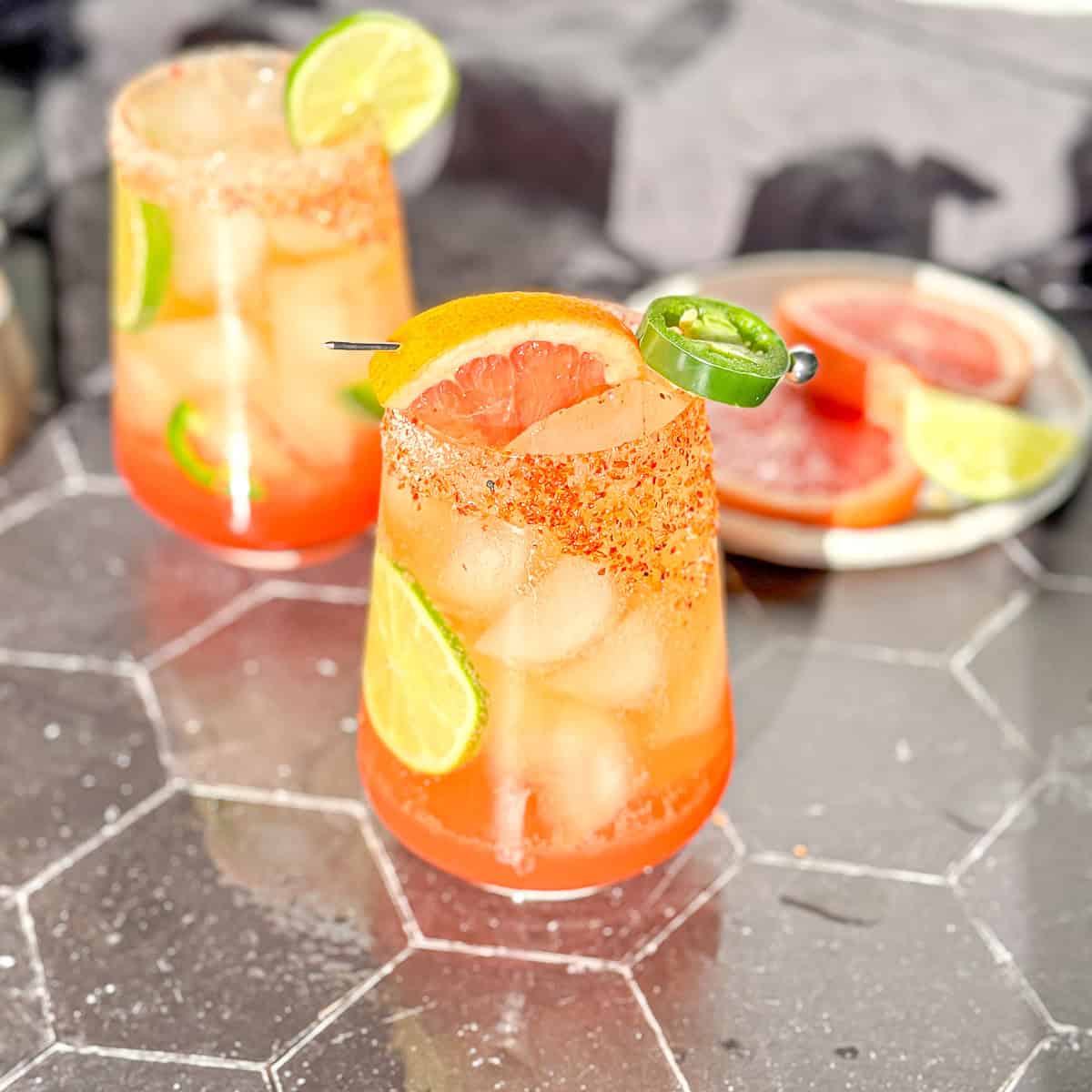 Photo of Two glasses of colorful cocktail garnished with lime and jalapeño slices on a hexagonal surface. | Wander Sober