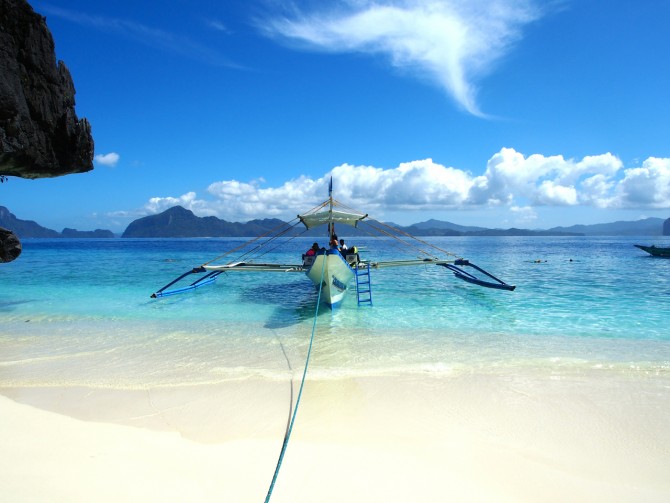 Philippines Island Hopping: Beaches, Water Sports, and Fresh Juices - My Sober Paradise Found in Crystal Waters and Vitamin Sea 5 Sover Travel - A traditional boat with outriggers is moored on a pristine white sandy beach with clear turquoise water. A blue sky with fluffy clouds stretches across the horizon, with distant mountains visible in the background.