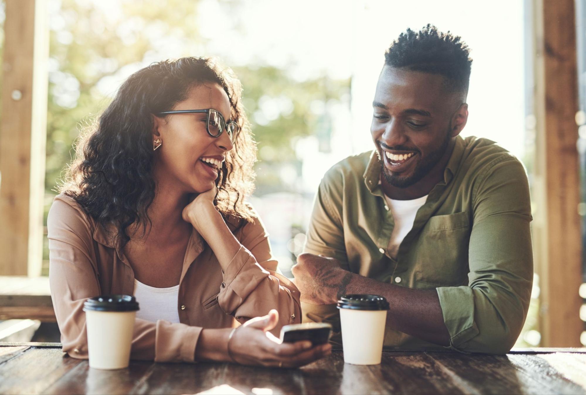 Sober Curious for Couples: Navigating a Healthier Relationship Together 5 Sover Travel - A woman and a man sit at an outdoor café table, smiling and looking at a smartphone in the womans hand. Both have cups of coffee in front of them. The setting is sunny and relaxed.