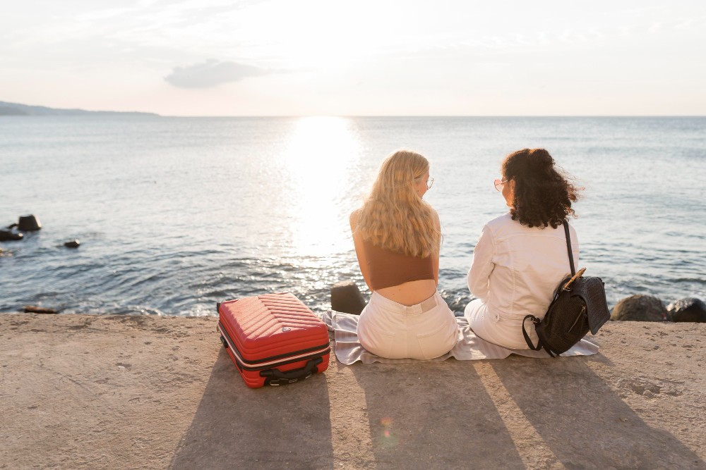 Sover Travel - Two people sit by the sea on a concrete surface at sunset, engrossed in their sober travel journey. Next to a red suitcase, they wear casual summer attire as the sun casts a golden reflection on the calm ocean, embodying a serene adventure of discovery.