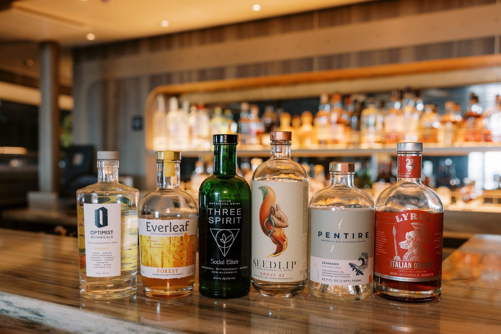 Sover Travel - Bottles of non-alcoholic spirits line the bar counter in a dimly lit setting, perfect for sober travelers. Each bottle boasts a distinct label and design, with shelves of various bottles blurred in the background, offering unique experiences at sea.