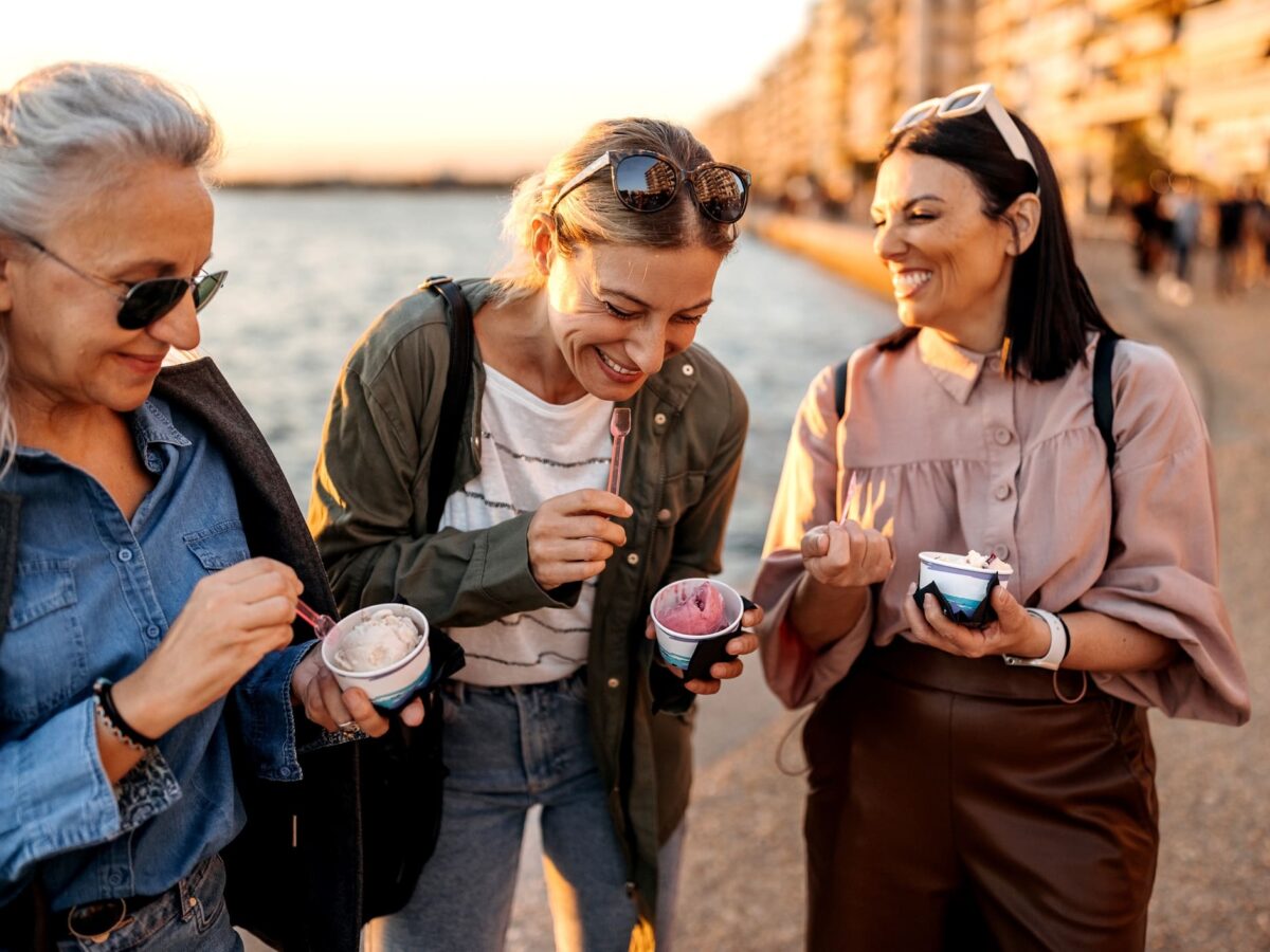 Traveling Sober: What to Expect and How to Embrace Every Journey 3 Sover Travel - Three women savor ice cream by the waterfront during sunset, embodying the spirit to Embrace Every Journey. Theyre smiling, dressed casually with sunglasses, as warm sunlight bathes the water on one side and buildings on the other.