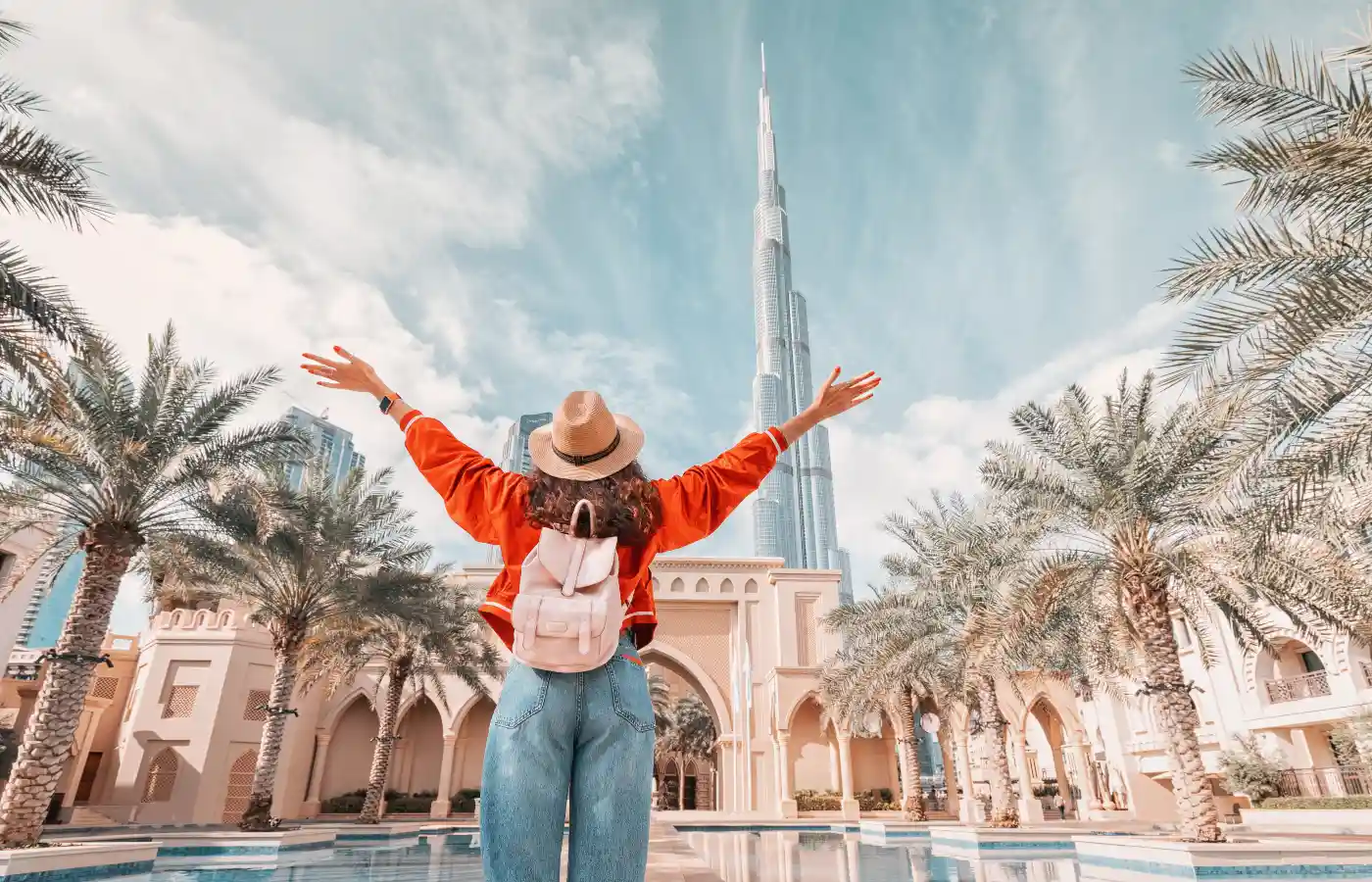 Woman with arms raised in joy, wearing a hat and backpack, in front of Burj Khalifa and palm trees, representing sober adventures in Dubai.