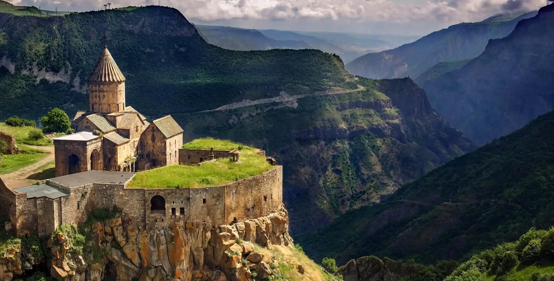 Photo of Tatev Monastery situated on a cliff in the mountains of Armenia with surrounding greenery. | Wander Sober
